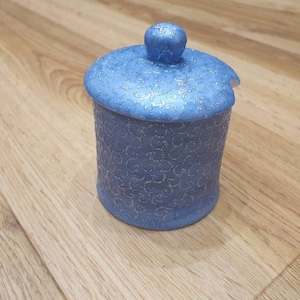 Puede incluir: Un tarro de cerámica azul con tapa y un pequeño pomo en la parte superior. El tarro y la tapa están decorados con un delicado patrón floral repetido y motas doradas. El tarro es cilíndrico y se asienta sobre una superficie de madera.