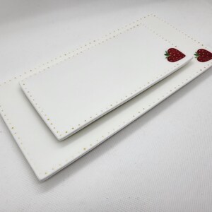 May include: Two white rectangular ceramic serving platters. The edges are decorated with small gold dots. Each platter features a red strawberry design. The platters are stacked, one on top of the other.