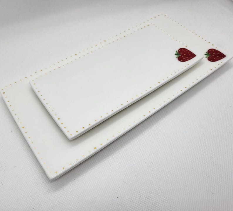 May include: Two white rectangular ceramic serving platters. The edges are decorated with small gold dots. Each platter features a red strawberry design. The platters are stacked, one on top of the other.