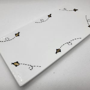 May include: A rectangular white ceramic serving dish with a glossy finish. The dish is decorated with a pattern of small yellow and black bees with dotted flight paths. The bees are arranged around the edges of the dish.