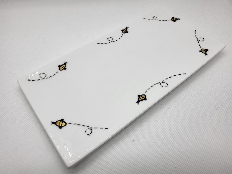 May include: A rectangular white ceramic serving dish with a glossy finish. The dish is decorated with a pattern of small yellow and black bees with dotted flight paths. The bees are arranged around the edges of the dish.