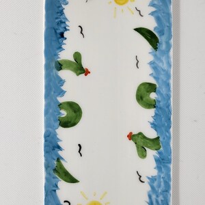 May include: A rectangular white ceramic tile featuring hand-painted blue borders on the left and right sides. The center displays two yellow suns, green cacti with small red accents, and several black bird silhouettes scattered across the surface.