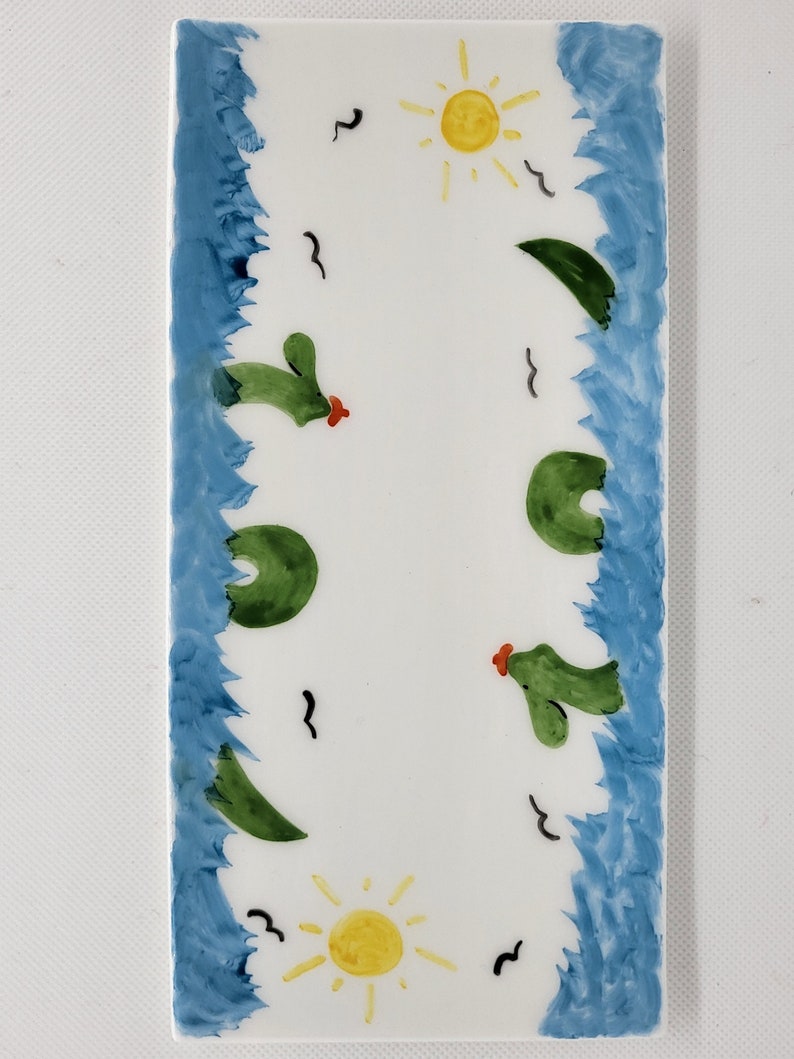 May include: A rectangular white ceramic tile featuring hand-painted blue borders on the left and right sides. The center displays two yellow suns, green cacti with small red accents, and several black bird silhouettes scattered across the surface.
