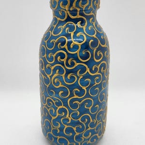 Peut inclure: Un vase en céramique bleu avec un motif tourbillonnant doré. Le vase a une forme cylindrique avec un dessus légèrement évasé. Le motif doré recouvre toute la surface du vase, créant un aspect décoratif et élégant.