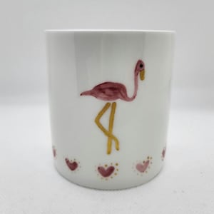 Puede incluir: Una taza de cerámica blanca con un flamenco rosa pintado a mano con largas patas doradas. La base de la taza está decorada con un patrón repetitivo de pequeños corazones rosas delineados con puntos dorados, creando un diseño limpio.