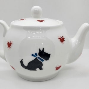 May include: White ceramic teapot featuring a black dog illustration with a blue bow. Red heart motifs decorate the teapot. It has a lid with a small knob and a curved handle. A charming kitchenware item.