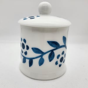 Puede incluir: Un tarro de cerámica blanco con tapa, adornado con un patrón floral azul. El tarro tiene una base redondeada y un pequeño pomo en la parte superior de la tapa. El diseño azul incluye flores y hojas estilizadas.