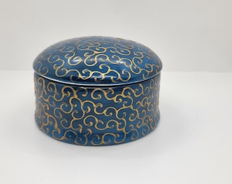 May include: A round, lidded ceramic box in a deep blue hue, decorated with a repeating gold swirl pattern. The box has a white rim and a matching gold design on the lid and sides. This decorative container is suitable for storing small items.