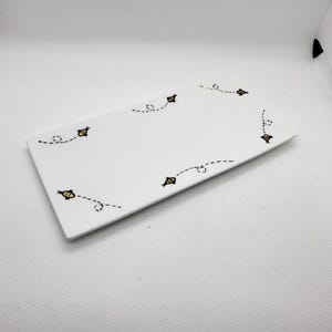 May include: A rectangular white ceramic serving tray with a whimsical bee design. The tray features several yellow and black bees with dotted flight paths, adding a playful touch. The tray is ideal for serving appetizers or desserts.
