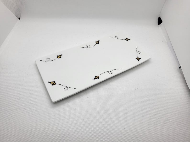 May include: A rectangular white ceramic serving tray with a whimsical bee design. The tray features several yellow and black bees with dotted flight paths, adding a playful touch. The tray is ideal for serving appetizers or desserts.