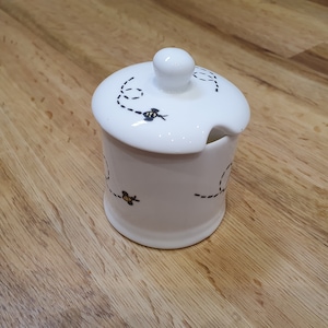 Può includere: Vasetto per miele in ceramica bianca con coperchio e intaglio per cucchiaio. Il vasetto è decorato con linee tratteggiate nere e api dei cartoni animati. Il coperchio ha un piccolo pomello per una facile manipolazione.