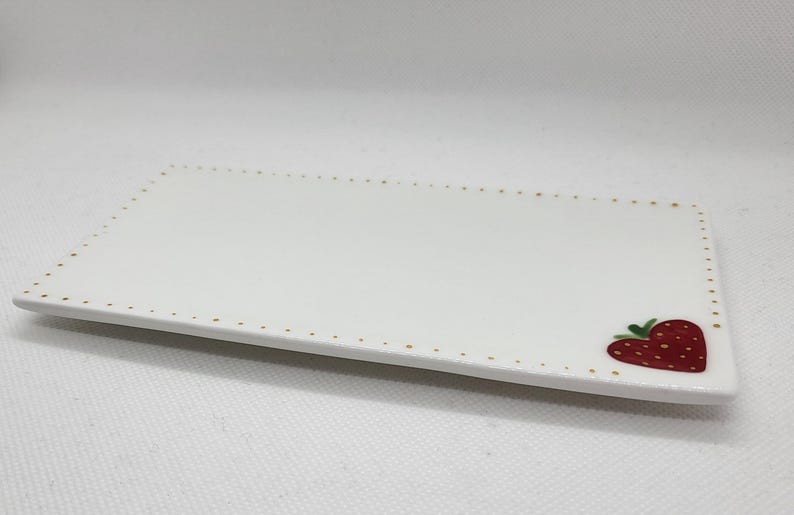 May include: A rectangular white ceramic serving platter with a strawberry design in the corner. The edge is decorated with small gold dots. The strawberry is red with green leaves.