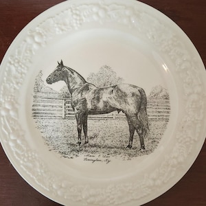 Może przedstawiać: Biały talerz ceramiczny z ozdobnym, wypukłym brzegiem. W centrum talerza znajduje się czarno-biała ilustracja konia na łące. Pod obrazkiem znajduje się napis "Man O' War Lexington, Ky".