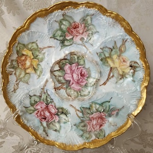 以下が含まれることがあります： 金色の縁と水色の背景を持つ、装飾的なスカラップエッジのプレート。 手描きのピンクと黄色のバラと緑の葉が特徴です。 透明なスタンドに飾られています。