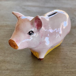 May include: A pink ceramic piggy bank with a yellow base and a slot on the back for coins. The piggy bank has a painted face with black eyes and a brown snout.