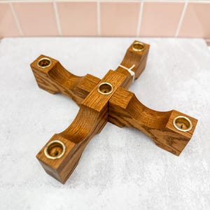 May include: A wooden candle holder with four arms, each holding a brass candle cup. The holder is made of light brown wood and has a cross-like design. The candle holder is a decorative item.