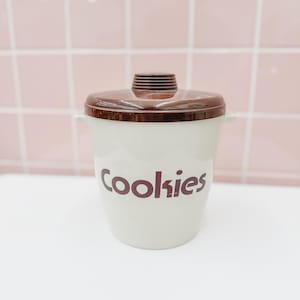 May include: A cream-colored ceramic cookie jar with a dark brown lid and a matching dark brown knob. The word Cookies is printed in dark brown lettering on the front of the jar. The container features two small side handles near the top rim.
