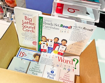 Pode incluir: Uma caixa de cartão cheia de materiais educativos. Inclui "Big Letter-Sound Cards", o currículo "Ready, Set, Read!" e vários pacotes para aprender fonética. A caixa também contém um cupom com 15% de desconto.