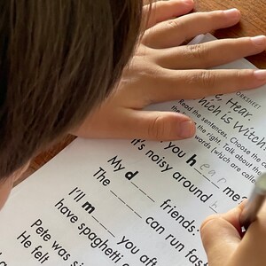 May include: A child is working on a worksheet with the title "Hear. Which is Witch?" The worksheet has sentences with missing letters. The child is writing the letter "h" in the sentence "It's noisy around you hear me?"