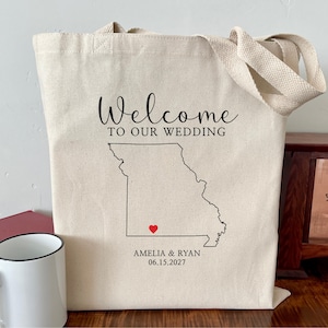May include: A beige canvas tote bag with the words "Welcome to Our Wedding" in black script. A black outline of the state of Missouri is below the text, with a red heart and the names Amelia & Ryan and the date 06.15.2027.