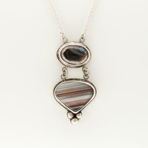 Puede incluir: Un collar con colgante de plata que presenta dos piedras apiladas engastadas en metal. La piedra superior es una ágata ovalada de bandas oscuras, y la inferior es una pieza más grande en forma de abanico con rayas horizontales marrones y blancas, terminada con tres pequeñas esferas de plata en la base.