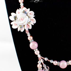 May include: A close-up of a necklace with a floral pendant and multiple strands of beads. The pendant is a white flower with pink accents. The necklace features pink and clear beads, with the text "DC Designs Cakes & Crafts".