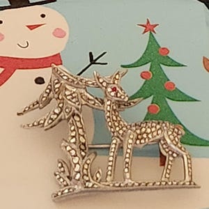 Könnte beinhalten: Silberfarbene Hirschbrosche mit roten Akzenten auf den Augen, neben einem stilisierten Baum. Die Brosche befindet sich vor einem hellblauen Hintergrund mit einer Schneemann- und Weihnachtsbaum-Illustration. Ein festliches Urlaubsaccessoire.