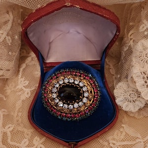 May include: A vintage brooch with a large, dark purple stone surrounded by a ring of clear rhinestones and a border of pink, blue, and green stones. The brooch is in a red velvet box with a blue velvet lining.