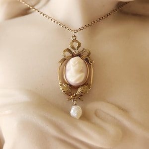 Può includere: Una collana con catena d'oro con un ciondolo a cammeo che presenta il profilo di una donna. Il cammeo è circondato da una cornice dorata con un fiocco e foglie. Una singola perla pende dalla parte inferiore del ciondolo.