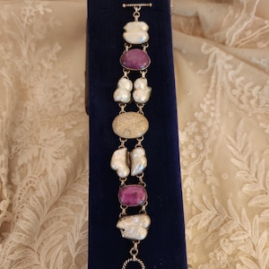 Könnte beinhalten: Ein silbernes Armband mit abwechselnd weißen, lila und beigefarbenen Steinen. Das Armband wird auf einem dunkelblauen Samtgrund präsentiert. Die Steine sind durch silberne Glieder verbunden, wodurch die Farben und Texturen hervorgehoben werden.