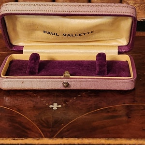 May include: A purple velvet-lined jewellery box with a gold clasp. The box is closed and has the words "PAUL VALLETTE" printed on the inside of the lid.