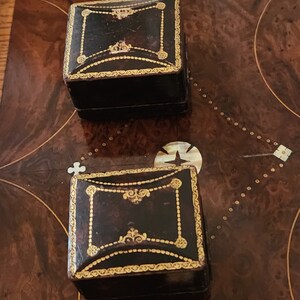 Puede incluir: Dos peque&ntilde;as cajas cuadradas de cuero negro con ribetes dorados y dise&ntilde;os ornamentados. Las cajas est&aacute;n sobre una superficie de madera.