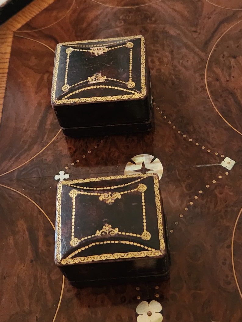 Puede incluir: Dos peque&ntilde;as cajas cuadradas de cuero negro con ribetes dorados y dise&ntilde;os ornamentados. Las cajas est&aacute;n sobre una superficie de madera.