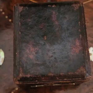 Puede incluir: Una peque&ntilde;a caja de cuero marr&oacute;n oscuro con una tapa con bisagras. La caja tiene un aspecto desgastado y vintage y probablemente se utiliza para guardar objetos peque&ntilde;os.