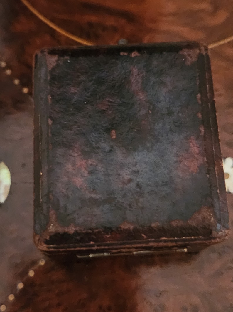 Puede incluir: Una peque&ntilde;a caja de cuero marr&oacute;n oscuro con una tapa con bisagras. La caja tiene un aspecto desgastado y vintage y probablemente se utiliza para guardar objetos peque&ntilde;os.