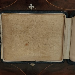 May include: A black velvet jewelry box with a beige interior. The box is open and the interior is empty.