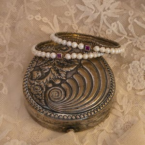 Pode incluir: Uma caixa de joias antiga prateada com detalhes florais e em espiral ornamentados. Duas pulseiras de pérolas com pequenos detalhes quadrados vermelhos repousam no topo. O fundo é um tecido de renda creme.