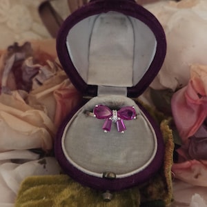 May include: A bow-shaped ring with pink gemstones and a small cluster of white stones in the center, set in a gold band. The ring is displayed inside a purple velvet ring box with a gray interior.