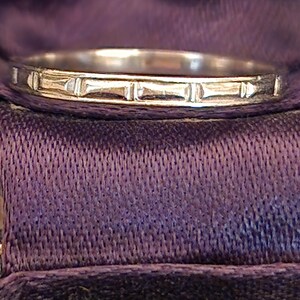 May include: A silver wedding band with a simple, textured design. The band is smooth and shiny, with a subtle pattern of lines.