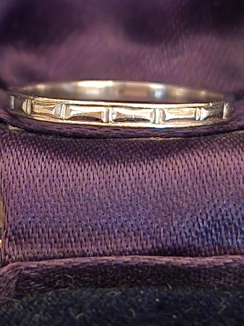 May include: A silver wedding band with a simple, textured design. The band is smooth and shiny, with a subtle pattern of lines.