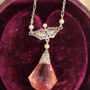 Puede incluir: Un delicado collar de plata con un colgante de cristal rosa y pequeños detalles de perlas. El collar se muestra en una caja forrada de terciopelo.