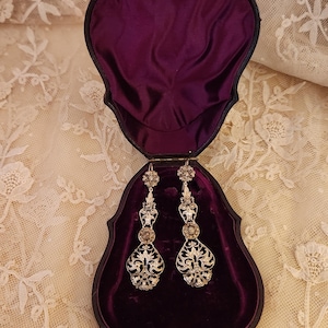 Könnte beinhalten: Ein Paar verzierter silberner Ohrringe, präsentiert in einer herzförmigen Vintage-Schmuckschatulle mit tiefviolettem Samt. Die Ohrringe weisen aufwendige Filigran- und Blumenmuster mit tropfenförmigem Anhänger auf.