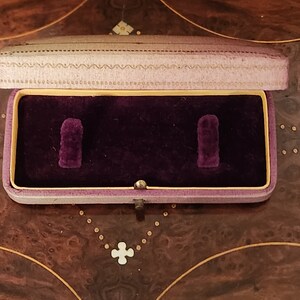 May include: A small, rectangular, purple velvet-lined jewellery box with a light purple lid. The box has two compartments and a gold-coloured latch.