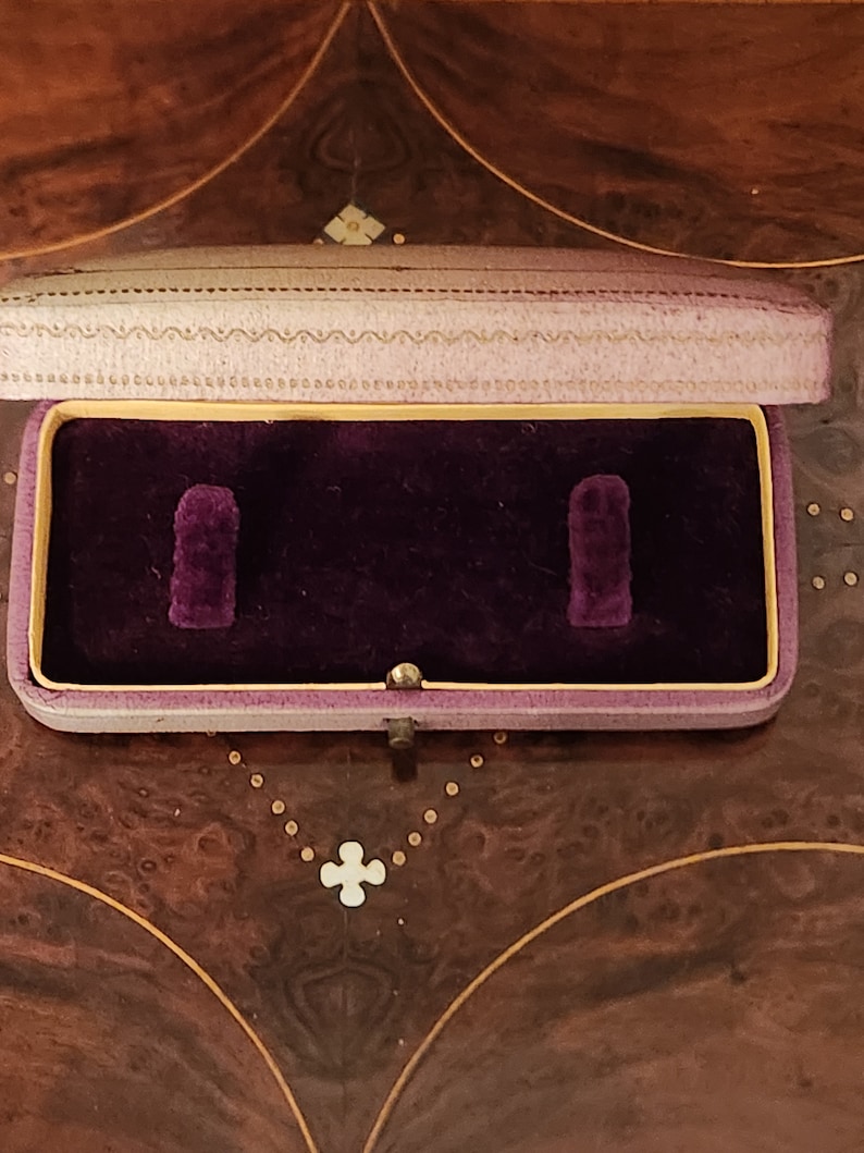 May include: A small, rectangular, purple velvet-lined jewellery box with a light purple lid. The box has two compartments and a gold-coloured latch.