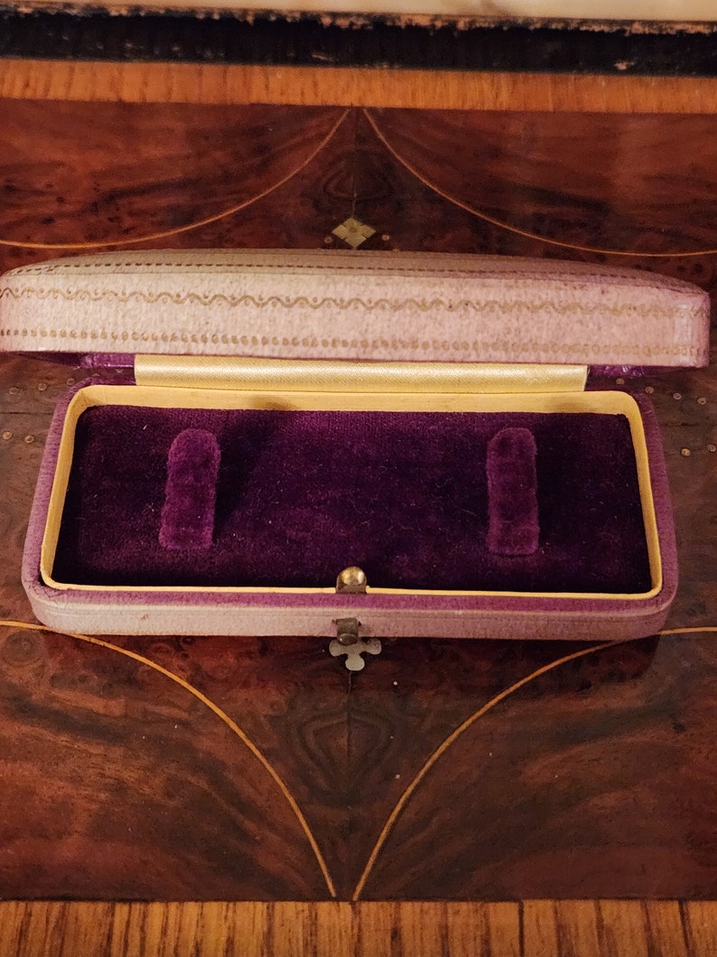 May include: A small, purple velvet jewellery box with a gold trim and a decorative pattern on the lid. The box is open and the interior is lined with purple velvet.