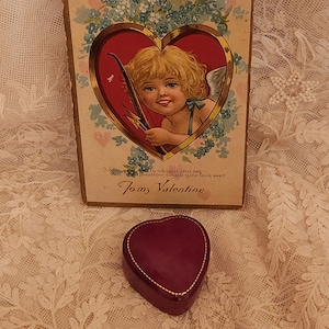May include: Vintage Valentine's Day card featuring a cherub with a bow and arrow, surrounded by blue flowers. The card reads "I love you truly as you can see, but precious is to do you love me?  Tomy Valentine". A small, heart-shaped, burgundy velvet box sits on a lace tablecloth.