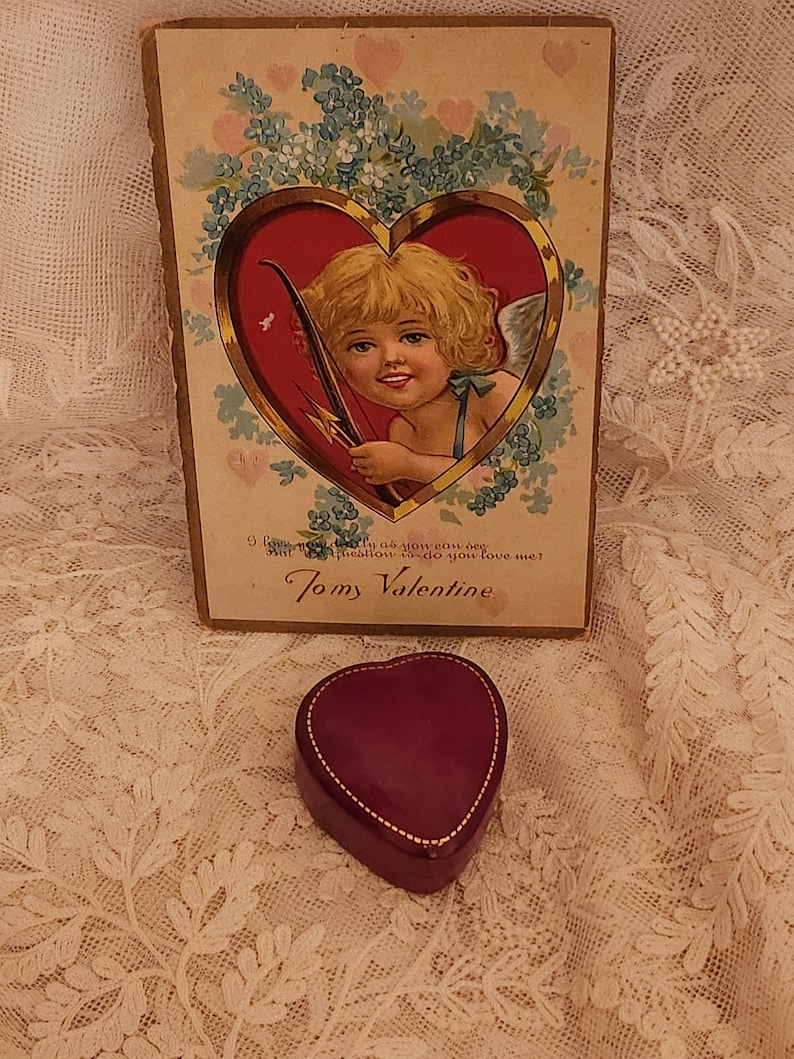 May include: Vintage Valentine's Day card featuring a cherub with a bow and arrow, surrounded by blue flowers. The card reads "I love you truly as you can see, but precious is to do you love me?  Tomy Valentine". A small, heart-shaped, burgundy velvet box sits on a lace tablecloth.