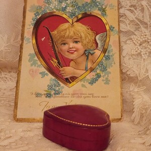 May include: Vintage Valentine's Day card featuring a cherub with a bow and arrow inside a heart. The card has a gold border and is surrounded by blue flowers. The text on the card reads "I love you dearly as you can see But the question is do you love me?"  A heart-shaped, burgundy velvet ring box sits on a lace tablecloth.