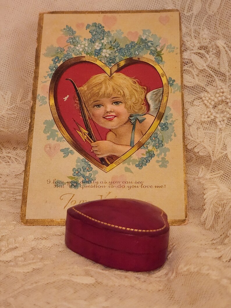 May include: Vintage Valentine's Day card featuring a cherub with a bow and arrow inside a heart. The card has a gold border and is surrounded by blue flowers. The text on the card reads "I love you dearly as you can see But the question is do you love me?"  A heart-shaped, burgundy velvet ring box sits on a lace tablecloth.