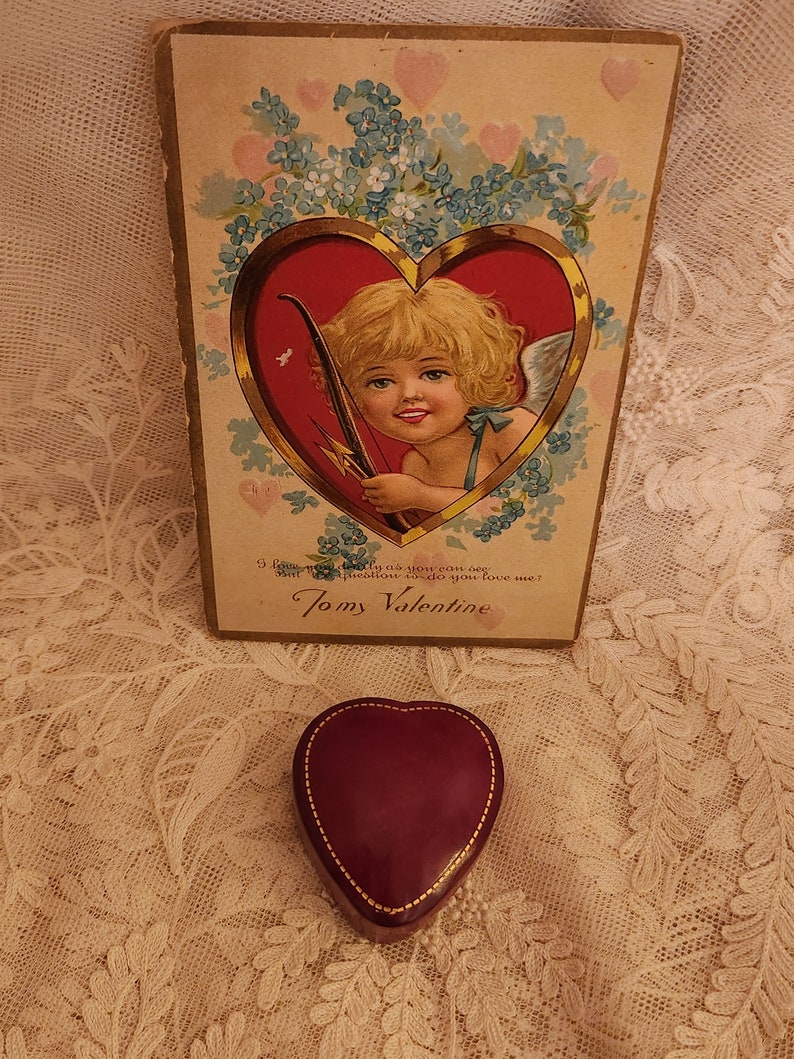 May include: Vintage Valentine's Day card featuring a cherub holding a bow and arrow, surrounded by blue flowers and hearts. The card reads "I love you as much as you can see, and I'll prove it, too, do you love me? To my Valentine."